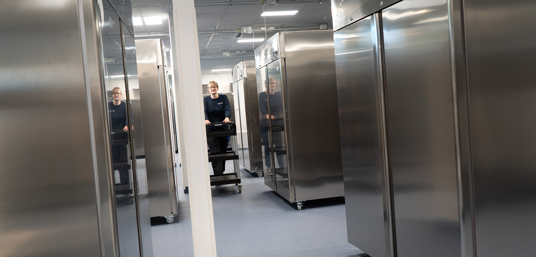 A person is pushing a wagon in a room filled with metal freezers. 
