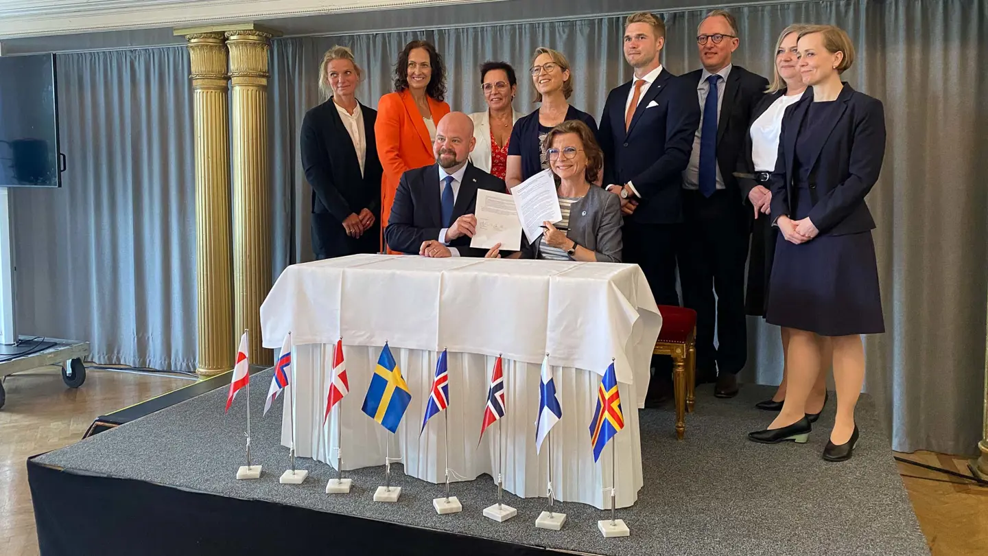 En grupp människor står runt ett vitt bord där en man och en kvinna håller upp ett underskrivet dokument.