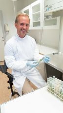 Man in lab coat sitting in a room holding a glass tube and smiling at the camera.