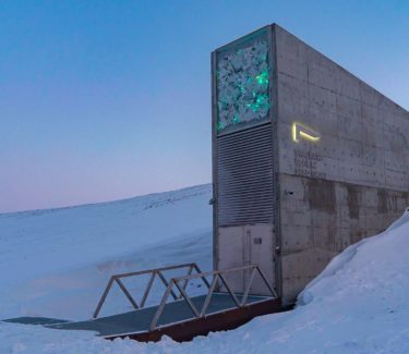 Exterior image of Svalbard Global Seed Vault surrounded by snow and with a sky of blue and pink in the background.