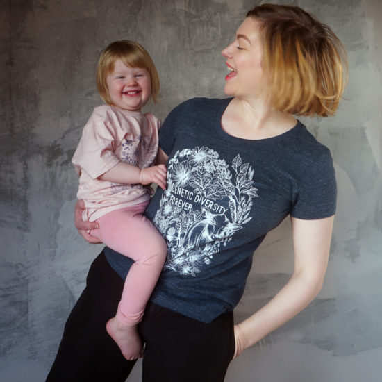 Kvinna med barn i famnen framför en betongvägg. Båda bär på t-shirt med tryck där det står Genetic Diversity Forever