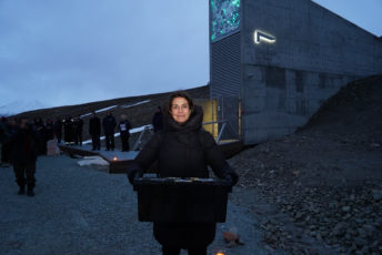 Woman in black coat standing in front of the Seed Vault holding a black seed box.