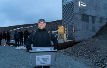 Man in black coat and a cap standing in front of the Seed Vault exterior holding a seed box.