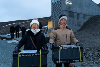 A woman wearing a black coat and white hat standing next to a man with beige jacket and white hat, both carrying seed boxes labeled Republic of Korea. The Seed Vault exterior in the background.